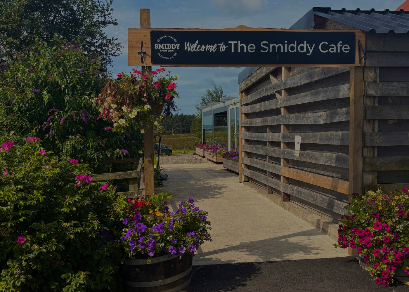 Entrance to The Smiddy Cafe with a wooden sign and flower beds on a sunny day.