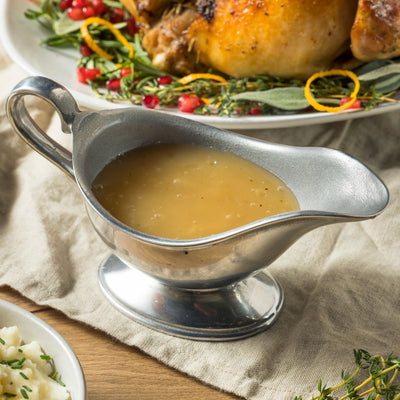 Metallic gravy boat filled with a rich on a wooden table with roast poultry in the background.